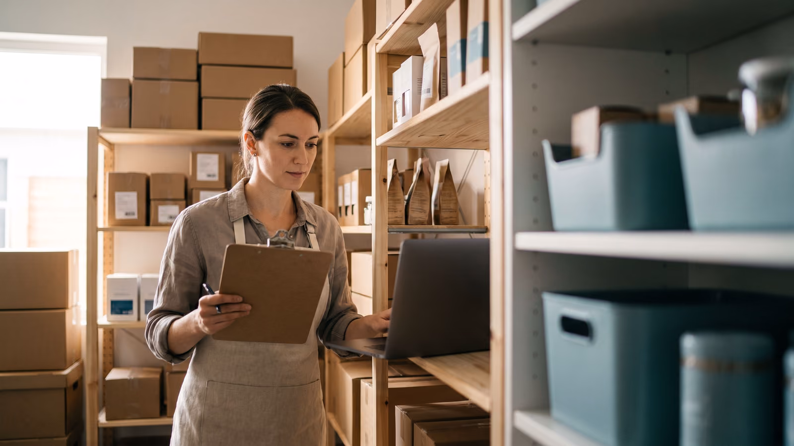 A shop owner checks a laptop next to shelves of boxed inventory in a small stockroom.