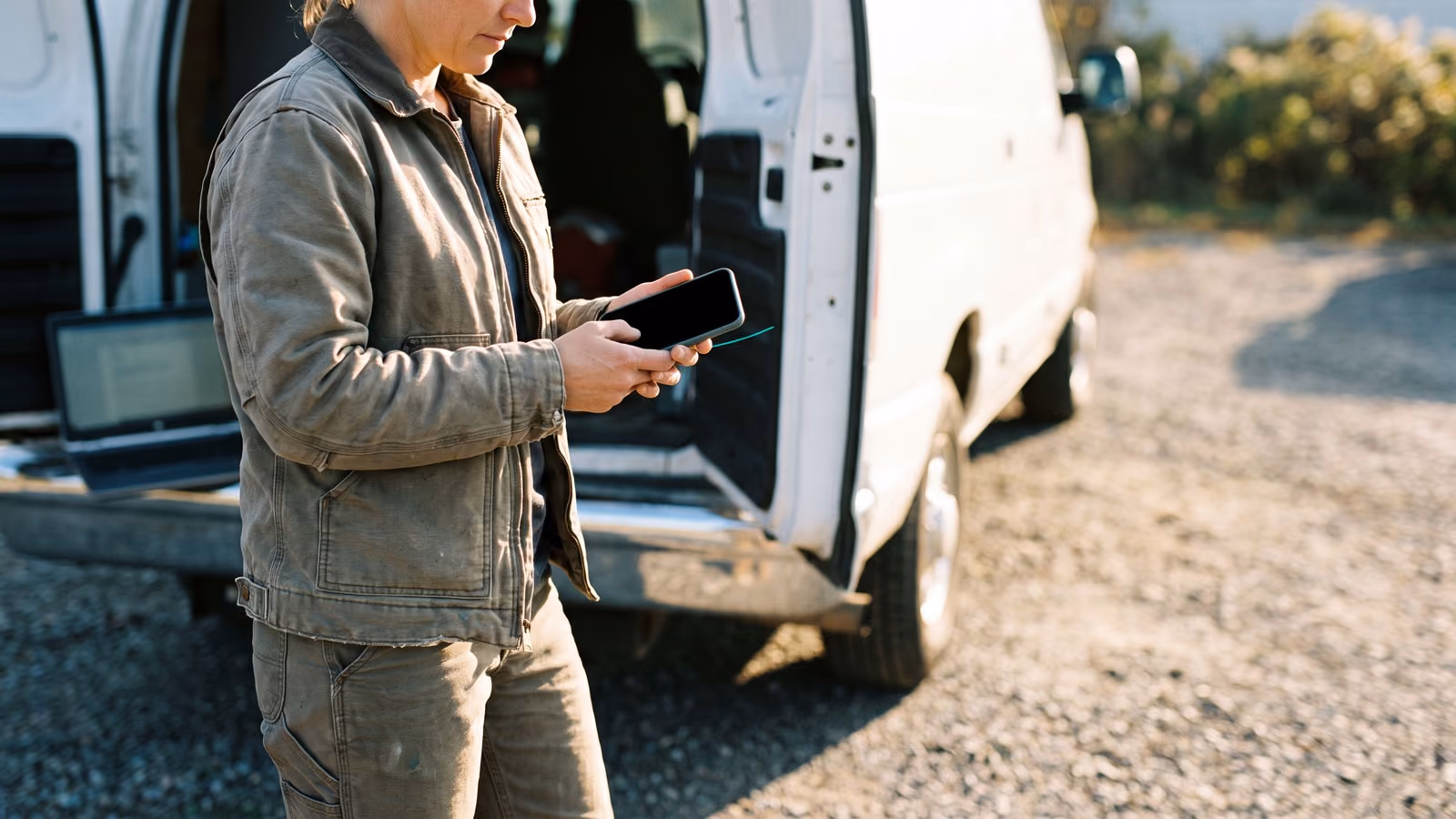 A tradesperson beside a service van checks a phone with a dark screen while a laptop sits open on the tailgate nearby.