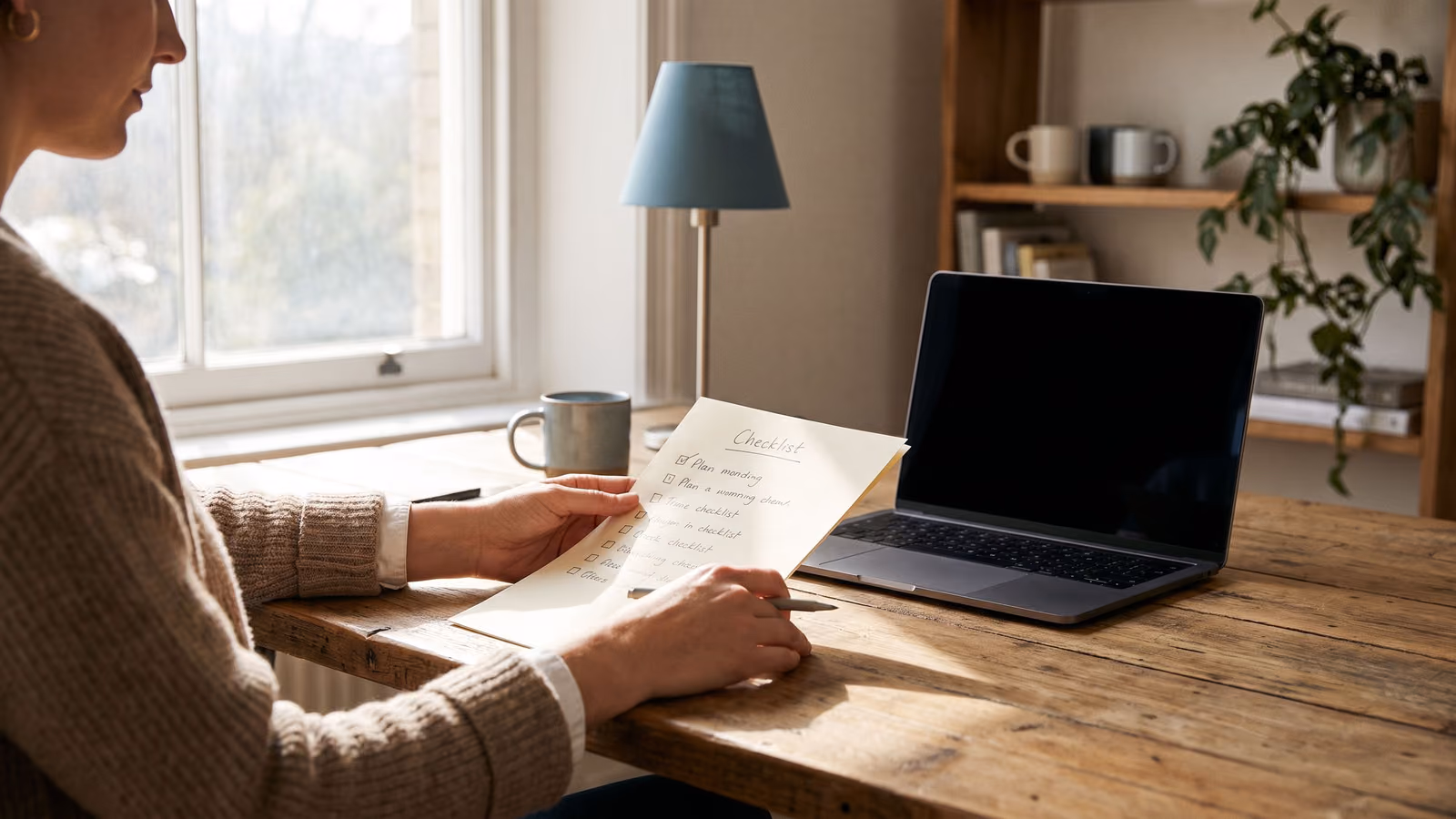 A person at a desk reviews a handwritten checklist next to a closed laptop in a softly lit office.