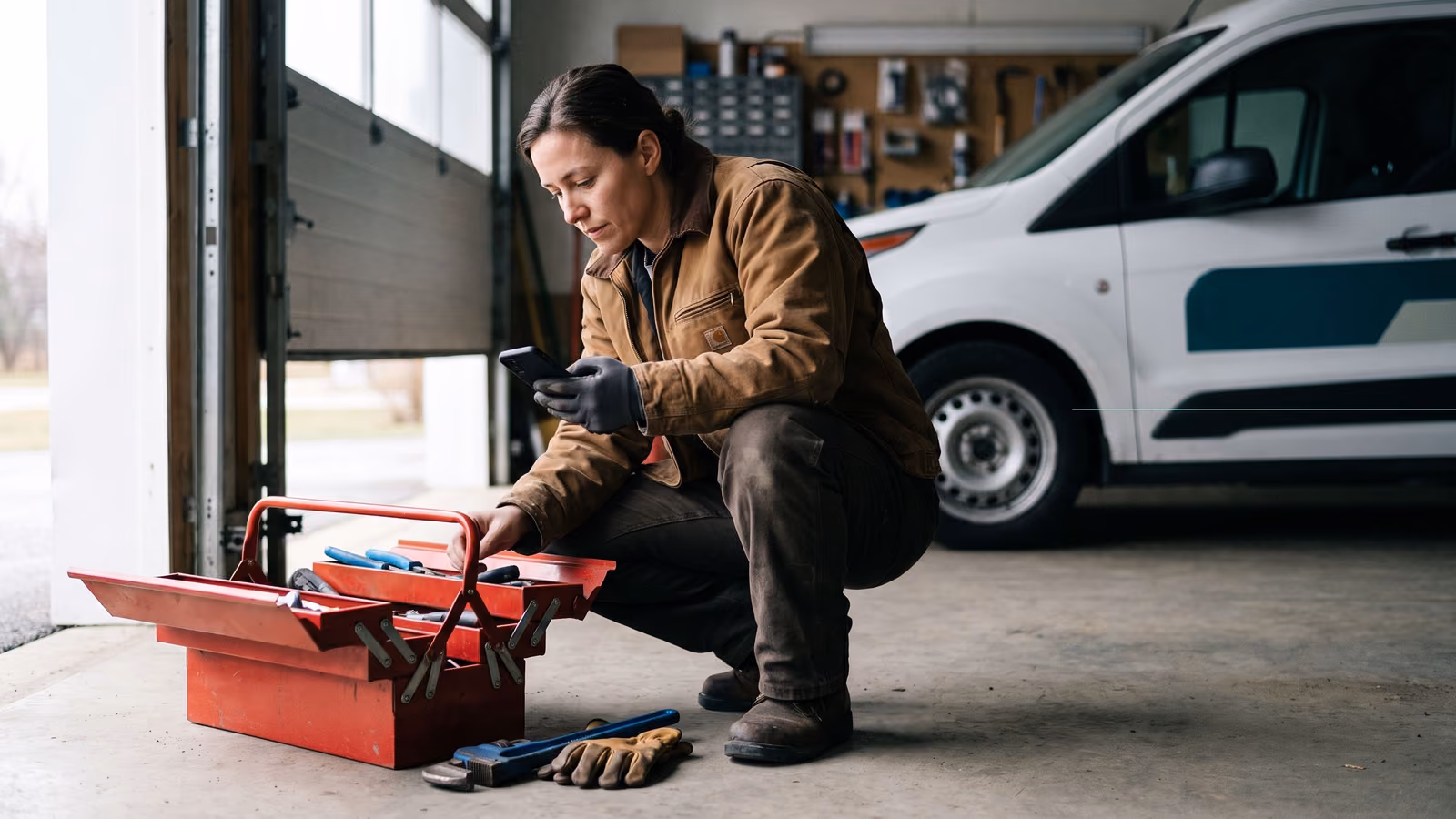 A tradesperson crouches by an open toolbox on a job site, checking a plain dark phone screen with a service van blurred in the background.