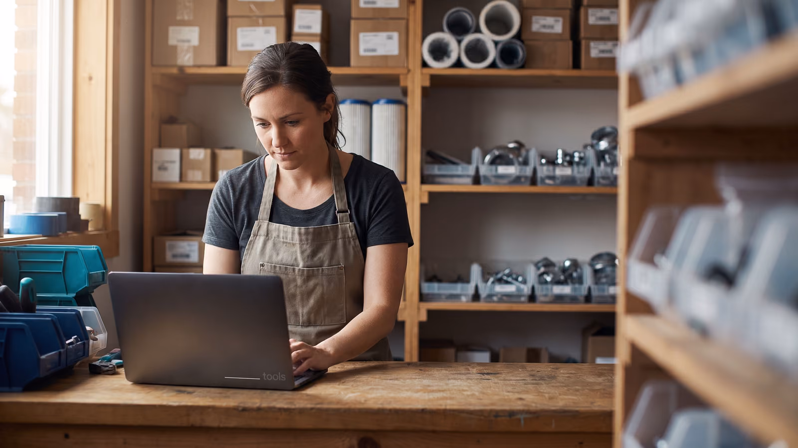 A woman in an apron looks at a laptop on a counter surrounded by shelves of parts and filters in a small trade storefront.