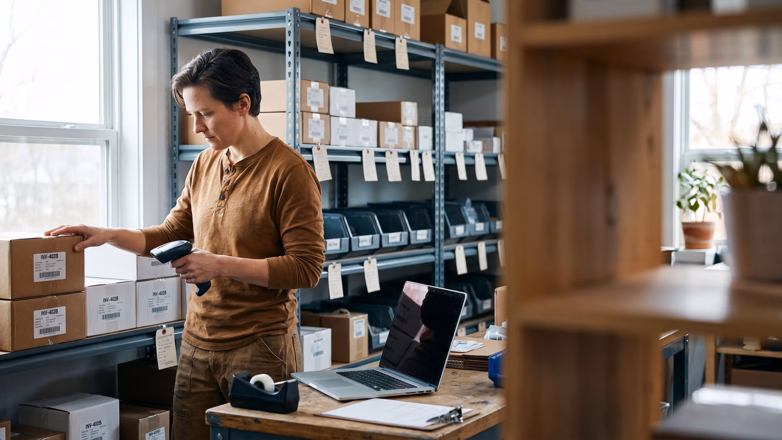A person in a stockroom checks a labeled parts box against a handheld scanner near an open laptop with a dark screen.