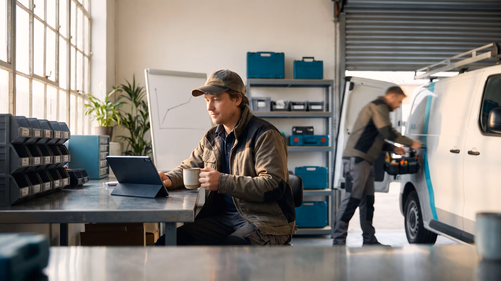 Business owner at a workbench calmly reading a tablet with coffee while a technician loads a van in the background.
